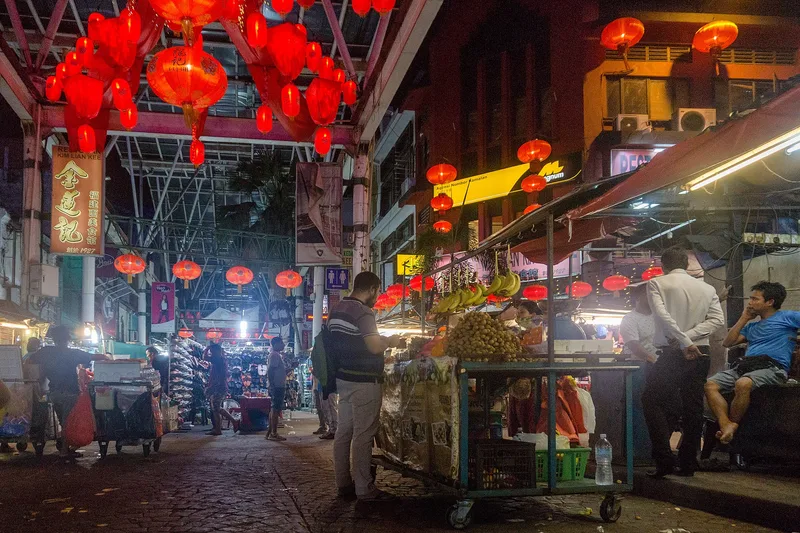 Petaling Street in Kuala Lumpur after dark, red lanterns strung overhead above rows of covered market stalls, with traders and shoppers around a fruit cart.