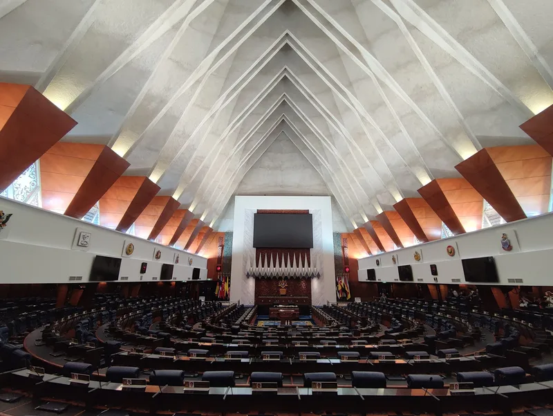 The empty debating chamber of the Dewan Rakyat, seen from the public gallery: tiers of vacant desks curving towards the Speaker's chair beneath the pinnacled white roof.