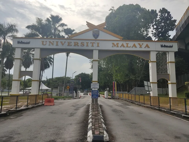 The main gate of Universiti Malaya in Kuala Lumpur, a white arched entrance lettered UNIVERSITI MALAYA beneath the university crest, with the empty campus road running through it.